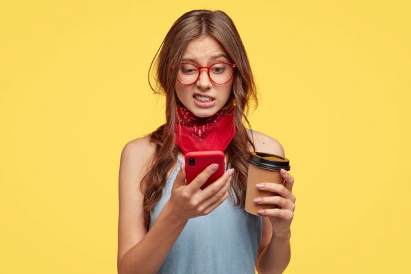 dissatisfied-lovely-woman-looks-with-disgust-and-aversion-at-cellular-edits-photo-in-special-app-drinks-coffee-to-go-poses-over-yellow-wall-feels-dislike-connected-to-high-speed-internet
